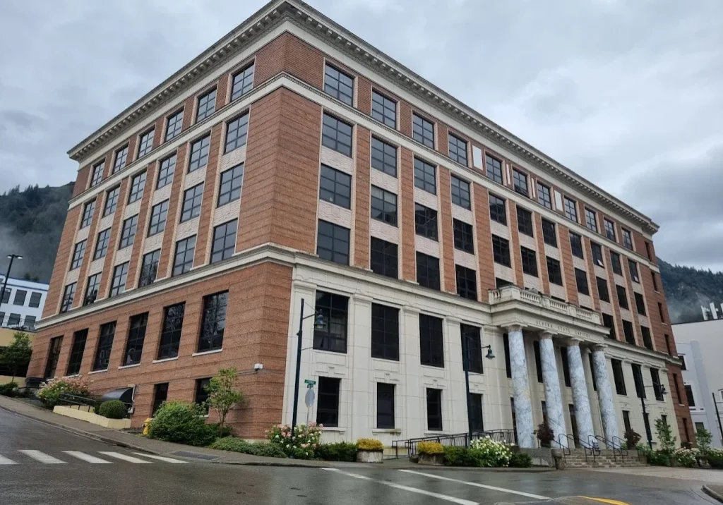 Three-story brick government building with a white stone entrance and six marble columns, labeled Alaska State Capitol, on a wet street under a cloudy sky, viewed from the corner.