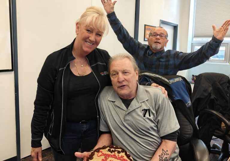 Smiling woman stands beside a man in a wheelchair who holds a round retirement cake that reads 'Congratulations on your retirement' in an office setting with two others raising their arms in the background.